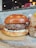 A juicy hamburger with a thick patty sitting on a slightly toasted bun. The burger features melted cheese beneath the patty, with a backdrop of a rustic interior with blurred lights and a cardboard container on the side.
