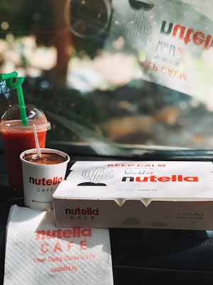 A takeaway setting featuring Nutella-themed items, including a branded cup filled with a chocolate spread and a takeaway box with a cactus illustration. Beside them, there is a cold beverage with a green straw and a napkin with Nutella Café branding. The ambient light suggests a warm and sunny environment.