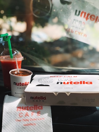 A takeaway setting featuring Nutella-themed items, including a branded cup filled with a chocolate spread and a takeaway box with a cactus illustration. Beside them, there is a cold beverage with a green straw and a napkin with Nutella Café branding. The ambient light suggests a warm and sunny environment.