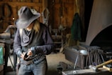 An older man wearing a wide-brimmed hat and leather clothing works intently in a workshop filled with various tools and equipment. He appears to be crafting or repairing an item, likely made of metal. The dimly lit room has a rugged, rustic feel with wooden walls and shelves laden with tools.