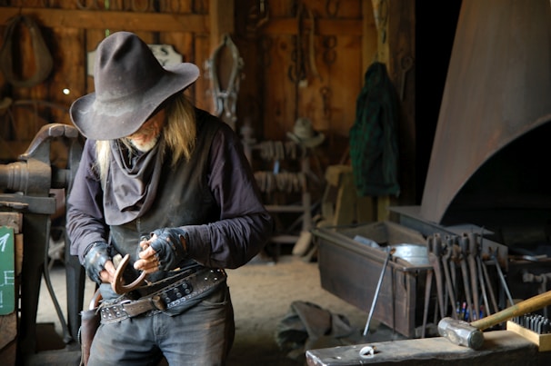 An older man wearing a wide-brimmed hat and leather clothing works intently in a workshop filled with various tools and equipment. He appears to be crafting or repairing an item, likely made of metal. The dimly lit room has a rugged, rustic feel with wooden walls and shelves laden with tools.