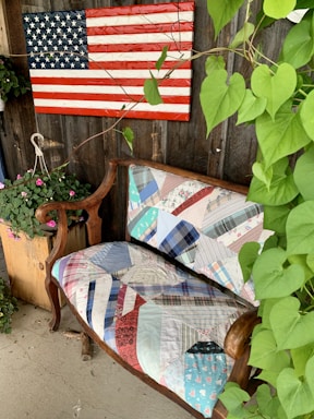 An American flag made of wooden planks is mounted on a rustic wooden wall. Below it, a vintage wooden bench is covered with a colorful, patchwork quilt. Green leafy vines and potted flowers add a touch of nature to the scene.