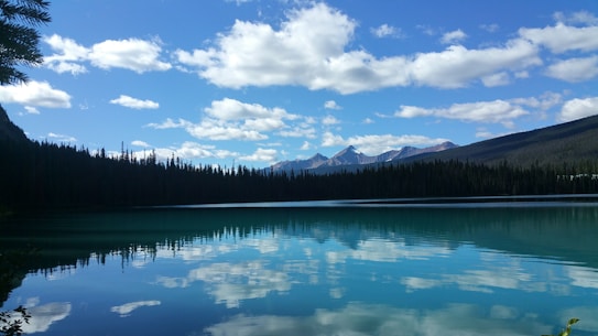 A serene lake with clear blue water reflecting the sky, clouds, and surrounding pine trees. Snow-capped mountains are visible in the distance under a sky dotted with fluffy white clouds. The landscape looks calm and peaceful.