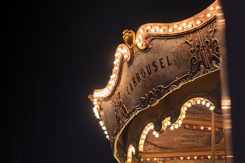 An illuminated carousel with ornate decorations and the word 'CARROUSEL' prominently displayed. The night setting enhances the warm glow of the numerous light bulbs adorning the structure's elegant curves and vintage floral patterns.