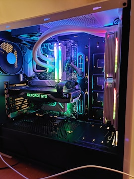 A well-organized and illuminated computer interior showcasing components like a GeForce GTX graphics card, illuminated RAM sticks, and other hardware. The setup features colorful LED lighting that enhances the aesthetic of the components and creates a vibrant visual appeal.