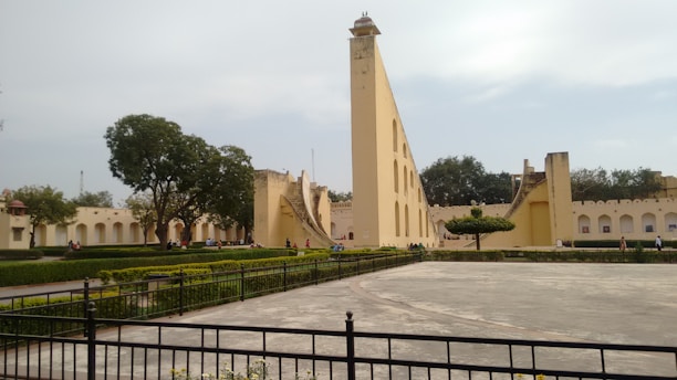 An ancient astronomical observatory with large beige stone structures and geometric designs, surrounded by green lawns and trees. People are walking and sitting along the pathways, and a metal fence borders the area in the foreground.