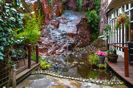 A serene garden setting features a small, tranquil pond surrounded by lush greenery and a rustic waterfall cascading over a rock face. The area is adorned with various potted plants and vibrant flowers. Wooden decking and seating provide a cozy atmosphere in this peaceful enclave adjacent to a building with large glass windows.