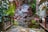A serene garden setting features a small, tranquil pond surrounded by lush greenery and a rustic waterfall cascading over a rock face. The area is adorned with various potted plants and vibrant flowers. Wooden decking and seating provide a cozy atmosphere in this peaceful enclave adjacent to a building with large glass windows.