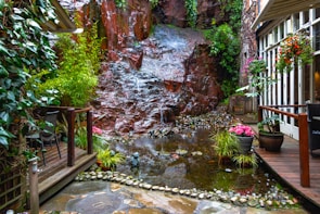 A serene garden setting features a small, tranquil pond surrounded by lush greenery and a rustic waterfall cascading over a rock face. The area is adorned with various potted plants and vibrant flowers. Wooden decking and seating provide a cozy atmosphere in this peaceful enclave adjacent to a building with large glass windows.