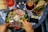 A hand is holding a fork, poised over a colorful and diverse salad in a black bowl, featuring prosciutto, avocado, roasted red peppers, olives, and sprinkled sesame seeds. Nearby are a glass of rose wine, a drink with a dried orange slice, a sandwich with leafy greens on whole grain bread, and a wallet with a gold credit card. The setting is on a decorative black and white tiled table.