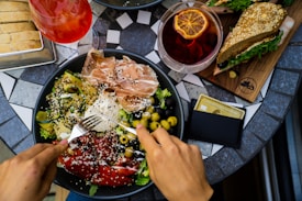 A hand is holding a fork, poised over a colorful and diverse salad in a black bowl, featuring prosciutto, avocado, roasted red peppers, olives, and sprinkled sesame seeds. Nearby are a glass of rose wine, a drink with a dried orange slice, a sandwich with leafy greens on whole grain bread, and a wallet with a gold credit card. The setting is on a decorative black and white tiled table.