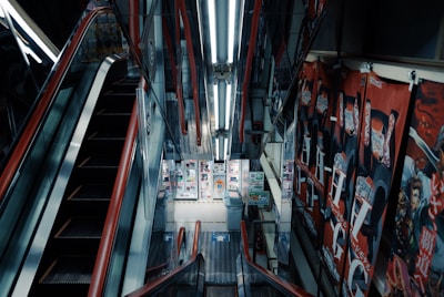 An escalator descends into a brightly lit underground area adorned with vertical advertising banners on the walls. The surrounding environment is enclosed, featuring metallic structures and linear fluorescent lighting overhead. A variety of colorful posters and small signs line the walls at the bottom of the escalator.