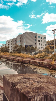 A modern apartment building complex is situated beside a narrow body of water. The sky is a vibrant blue with scattered clouds, and a wooden structure is visible in the foreground. Trees and greenery surround the buildings, reflecting softly in the water.