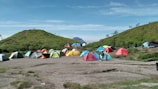 A large group of colorful tents is scattered across a grassy campsite with a mountainous backdrop. Two hills frame the campsite, covered with green vegetation. A few people are walking around the tents, suggesting an outdoor gathering or camping trip. The sky is clear and blue, enhancing the peaceful outdoor setting.
