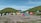 A large group of colorful tents is scattered across a grassy campsite with a mountainous backdrop. Two hills frame the campsite, covered with green vegetation. A few people are walking around the tents, suggesting an outdoor gathering or camping trip. The sky is clear and blue, enhancing the peaceful outdoor setting.