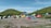 A large group of colorful tents is scattered across a grassy campsite with a mountainous backdrop. Two hills frame the campsite, covered with green vegetation. A few people are walking around the tents, suggesting an outdoor gathering or camping trip. The sky is clear and blue, enhancing the peaceful outdoor setting.