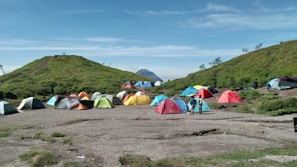 A large group of colorful tents is scattered across a grassy campsite with a mountainous backdrop. Two hills frame the campsite, covered with green vegetation. A few people are walking around the tents, suggesting an outdoor gathering or camping trip. The sky is clear and blue, enhancing the peaceful outdoor setting.