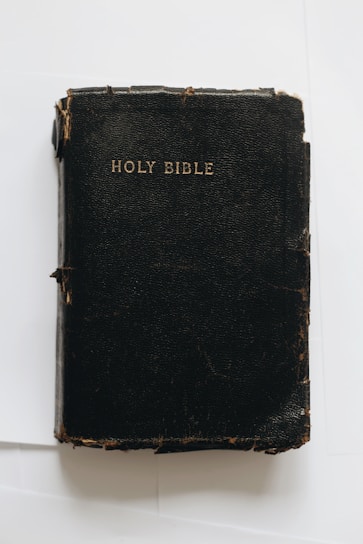 A worn, black leather-bound book with the title 'Holy Bible' embossed in gold on the cover. The book appears to show signs of aging, with frayed edges and a slightly cracked surface.