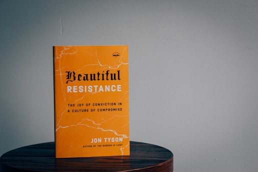 A book with a bright orange cover resting on a wooden surface. The title 'Beautiful Resistance' is prominently displayed in bold, stylized font, and the author's name, Jon Tyson, is indicated. The background is plain with a subtle, muted tone.
