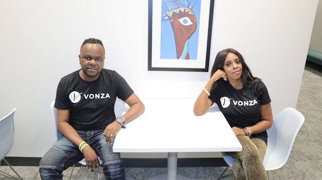 Two individuals sit at a small white table in a modern setting, both wearing black shirts with the logo 'VONZA.' They seem relaxed and are sitting on white chairs. A framed abstract artwork is displayed on the wall behind them.