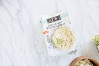 A package of Alexia White Cheddar Riced Cauliflower with Black Pepper resting on a marble surface. The packaging is primarily white and green, and shows an image of the riced cauliflower dish. To the right of the package, there is a wooden bowl containing the same dish.