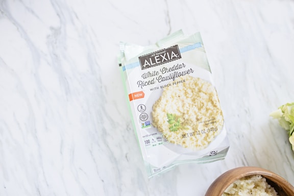 A package of Alexia White Cheddar Riced Cauliflower with Black Pepper resting on a marble surface. The packaging is primarily white and green, and shows an image of the riced cauliflower dish. To the right of the package, there is a wooden bowl containing the same dish.