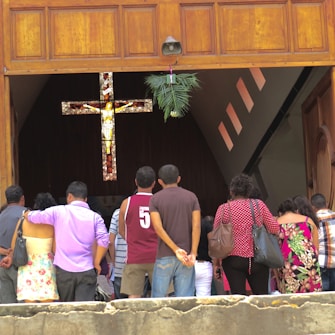 A group of people stand in front of a wooden doorway leading into a dimly lit space, possibly a church, with a stained glass cross visible inside. The doorway has decorations with greenery hanging above.