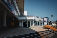 A corner art store surrounded by storefronts displaying various posters and artwork with several signs and posters in the windows. The building has a modern architectural design with large glass windows. A poster stand nearby advertises 'Freedom of Expression' with vibrant colors contrasting against the neutral tones of the building. Sunlight casts sharp shadows on the concrete pavement.
