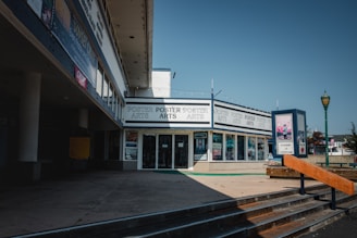 A corner art store surrounded by storefronts displaying various posters and artwork with several signs and posters in the windows. The building has a modern architectural design with large glass windows. A poster stand nearby advertises 'Freedom of Expression' with vibrant colors contrasting against the neutral tones of the building. Sunlight casts sharp shadows on the concrete pavement.