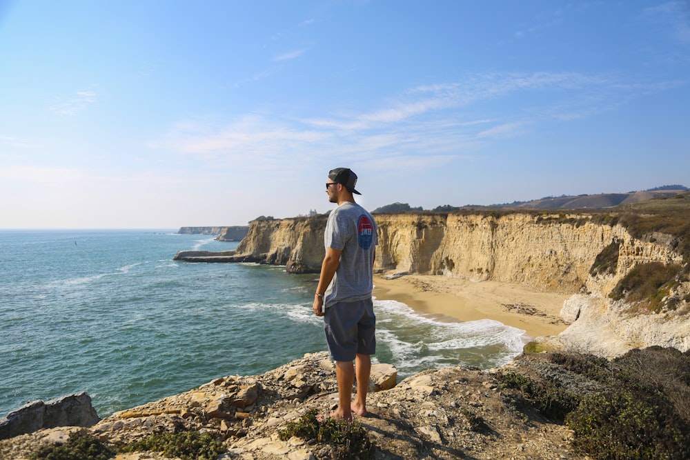 man in white t-shirt standing on brown rock formation near body of water during daytime