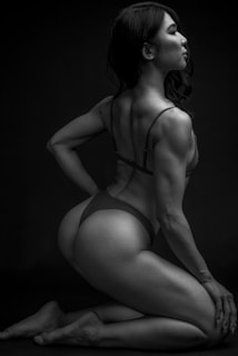 A black and white photograph features a person with long hair and a muscular build posing on their knees. The lighting highlights the contours and definition of their muscles, creating strong contrasts against the dark background. The person is wearing minimal clothing, and their posture suggests confidence and strength.
