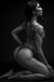 A black and white photograph features a person with long hair and a muscular build posing on their knees. The lighting highlights the contours and definition of their muscles, creating strong contrasts against the dark background. The person is wearing minimal clothing, and their posture suggests confidence and strength.