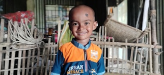 A young child with a shaved head, wearing an orange and blue shirt, stands smiling in front of a bamboo and wooden structure. The background is cluttered with various household items including ropes and baskets.