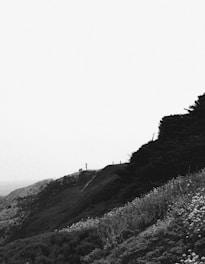 A black and white landscape photograph captures a lone figure standing on a high cliffside. The scene includes rugged terrain with patches of vegetation covering the hillside. A vast expanse of sky dominates the upper portion of the image, creating a sense of openness and emptiness.