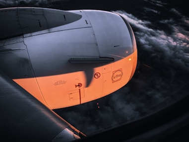 A close-up view of an airplane engine, partially illuminated by a warm, orange sunset light. The body of the engine is white with technical markings and logos visible. Surrounding the engine, dark clouds create a dramatic background, indicating the aircraft is flying at a high altitude.