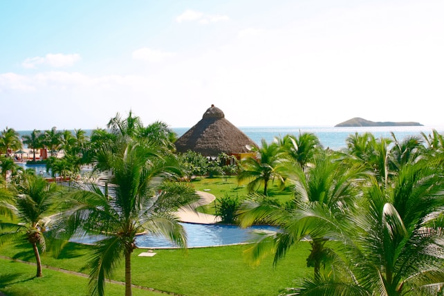 A tropical resort setting with lush green palm trees surrounding a pool area. A thatched-roof hut is visible in the center, with the serene ocean and small islands in the background under a clear blue sky.