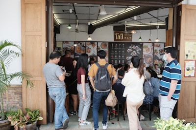 A group of people is gathered outside a restaurant, waiting to enter. The restaurant has an open entrance with wooden doors and displays menu items on the walls inside. Some people are casually dressed and a few are wearing masks, indicating a casual dining environment.