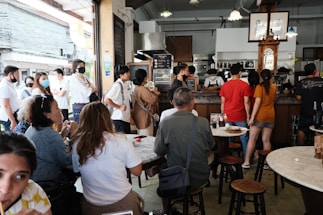 A crowded cafe or restaurant scene with numerous people standing in line and sitting at tables. Customers are engaging in conversation, some wearing face masks. The interior features wooden furniture and warm lighting, with a counter at the back where staff are serving drinks and food.