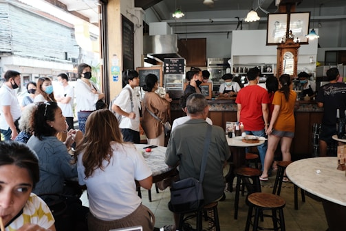 A crowded cafe or restaurant scene with numerous people standing in line and sitting at tables. Customers are engaging in conversation, some wearing face masks. The interior features wooden furniture and warm lighting, with a counter at the back where staff are serving drinks and food.