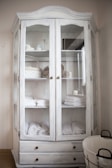 A white wooden cabinet with glass doors is filled with neatly arranged folded towels, rolled bath linens, and a few decorative items like baskets and containers. Two lower drawers are visible, and to the side, there is a round wire basket on the floor.