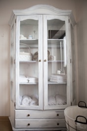 A white wooden cabinet with glass doors is filled with neatly arranged folded towels, rolled bath linens, and a few decorative items like baskets and containers. Two lower drawers are visible, and to the side, there is a round wire basket on the floor.