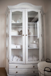 A white wooden cabinet with glass doors is filled with neatly arranged folded towels, rolled bath linens, and a few decorative items like baskets and containers. Two lower drawers are visible, and to the side, there is a round wire basket on the floor.