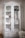 A white wooden cabinet with glass doors is filled with neatly arranged folded towels, rolled bath linens, and a few decorative items like baskets and containers. Two lower drawers are visible, and to the side, there is a round wire basket on the floor.
