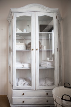 A white wooden cabinet with glass doors is filled with neatly arranged folded towels, rolled bath linens, and a few decorative items like baskets and containers. Two lower drawers are visible, and to the side, there is a round wire basket on the floor.