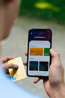 A person holding a smartphone displaying a colorful app interface with categories such as 'Supermarkt' and 'Opticien'. The person is also holding a credit card in the other hand.