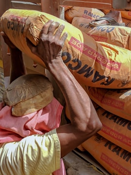 A person is carrying a heavy bag labeled 'RAMCO' over their shoulder. The person is wearing a head covering and a pink cloth around the neck while loading bags into a vehicle.