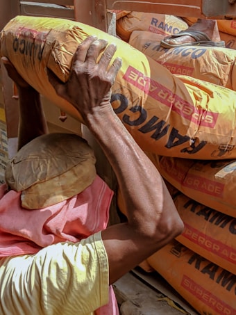 A person is carrying a heavy bag labeled 'RAMCO' over their shoulder. The person is wearing a head covering and a pink cloth around the neck while loading bags into a vehicle.