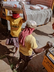 Two workers are carrying heavy bags of construction material, possibly cement, on their heads. They are wearing yellow shirts and have their faces covered with red cloths, likely for protection from dust or the sun. The scene takes place near a truck loaded with similar bags, indicating manual labor and transportation of goods.