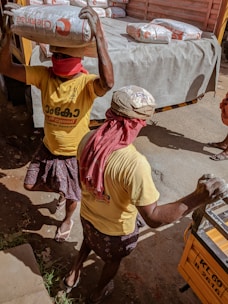 Two workers are carrying heavy bags of construction material, possibly cement, on their heads. They are wearing yellow shirts and have their faces covered with red cloths, likely for protection from dust or the sun. The scene takes place near a truck loaded with similar bags, indicating manual labor and transportation of goods.