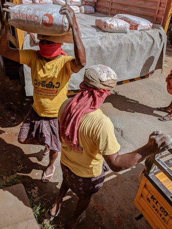 Two workers are carrying heavy bags of construction material, possibly cement, on their heads. They are wearing yellow shirts and have their faces covered with red cloths, likely for protection from dust or the sun. The scene takes place near a truck loaded with similar bags, indicating manual labor and transportation of goods.